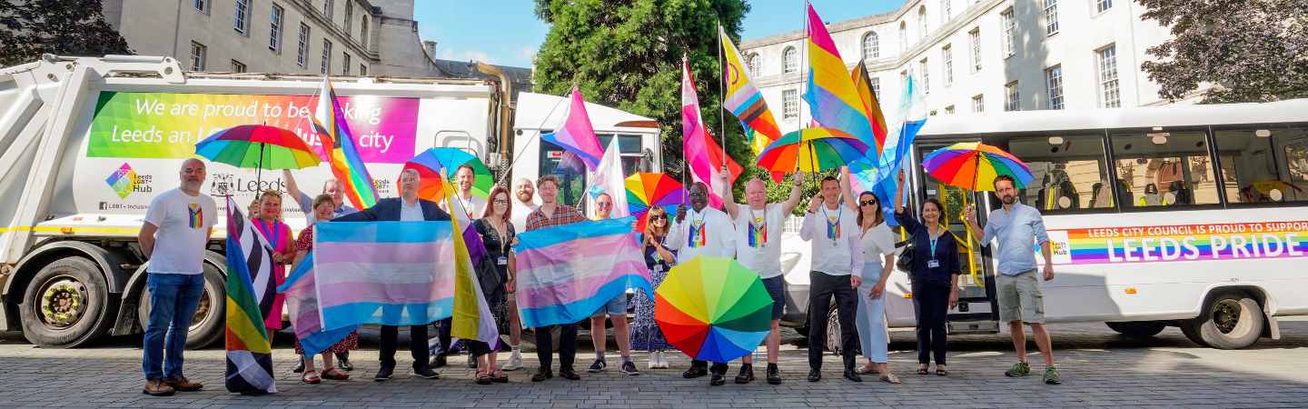 A group of people are waving rainbow coloured flags and umbrellas as they stand in front of two Leeds City Council vehicles which display messages of support for Leeds Pride