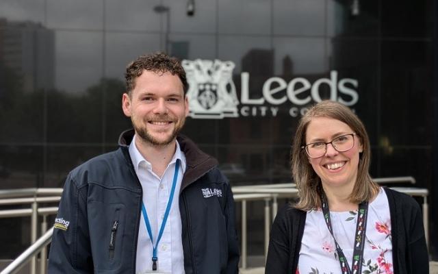 A man and a woman stand smiling outside their place of work. They are engineers at Leeds City Council and in the background is the council logo in white on a shiny black wall.