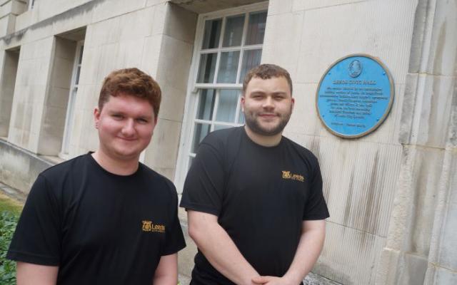Two employees wearing Leeds City Council branded T-shirts are smiling outside the Civic Hall with a blue plaque in the background 